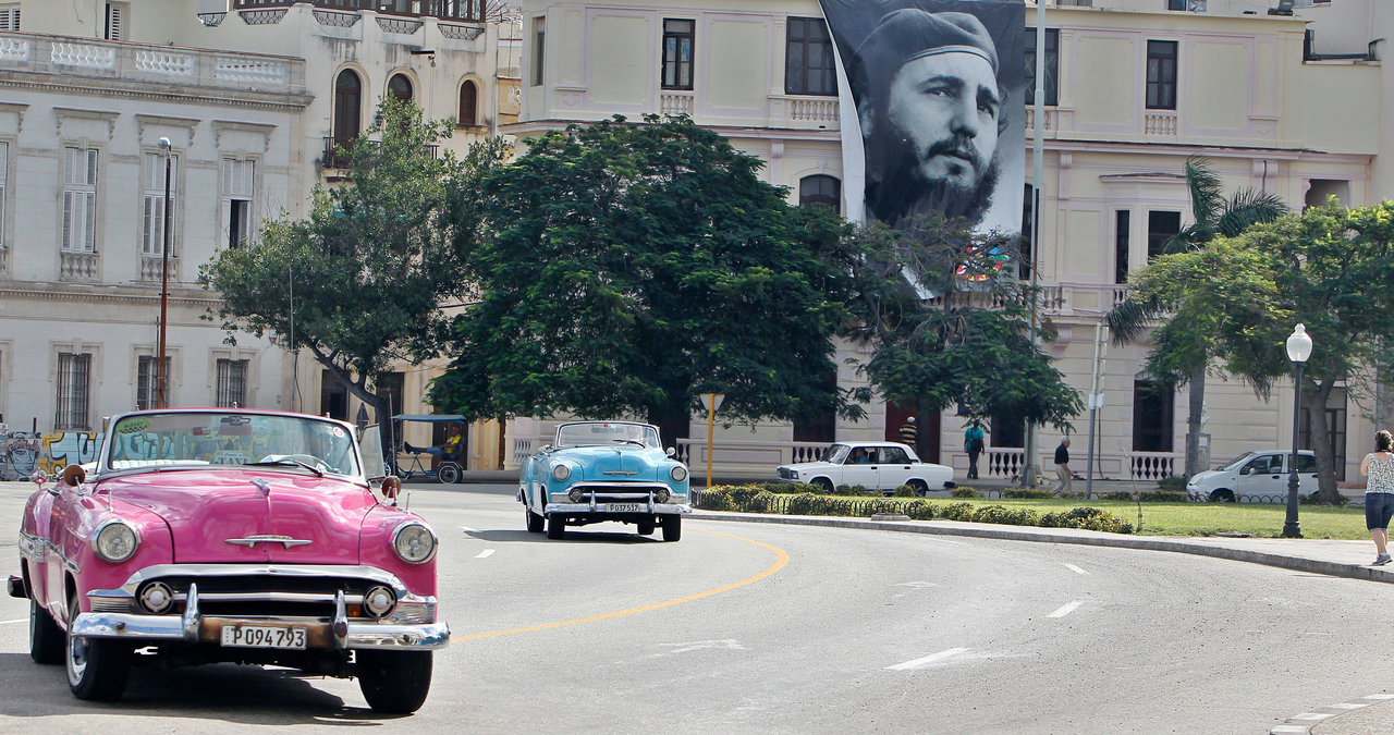 Dos viejos automóviles pasan junto a un edificio donde se expone una imagen de Fidel Castro.