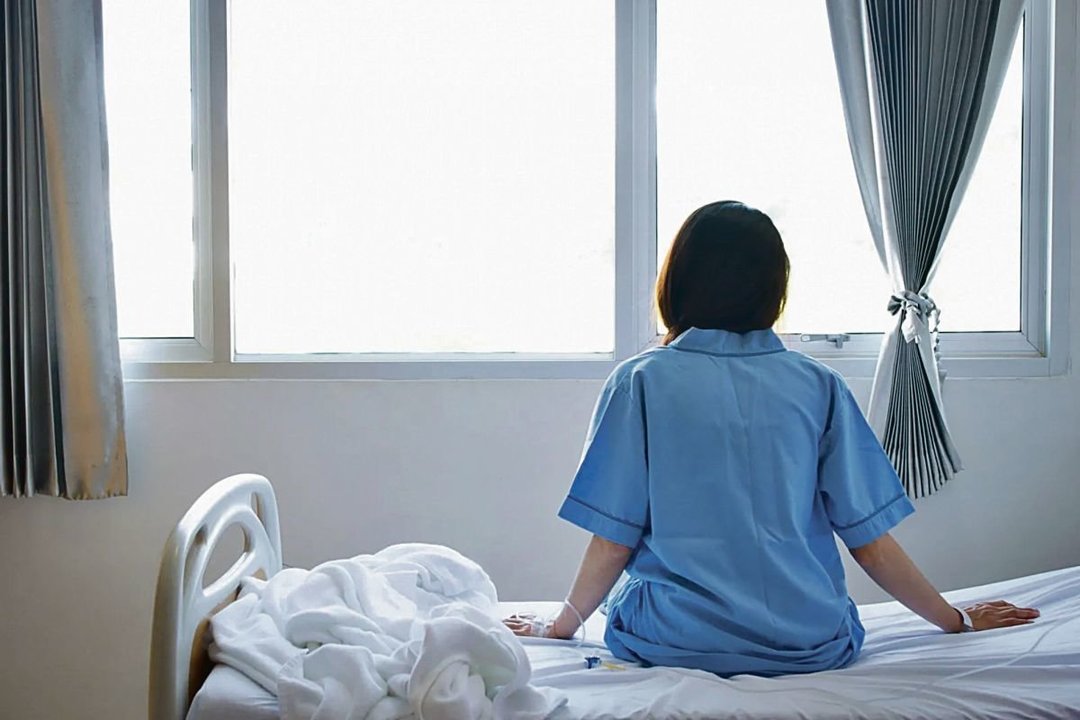 Una mujer joven, sentada sobre la cama de su habitación de hospital al lado de una ventana.