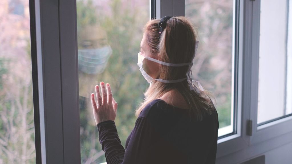 Una mujer protegida con una mascarilla mira por la ventana de su casa.