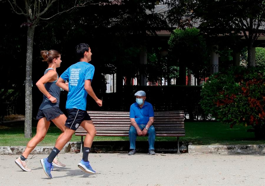 Un hombre con mascarilla descansa en un banco mientras una pareja de corredores practica deporte en el Parque de Rosalía de Castro en Lugo