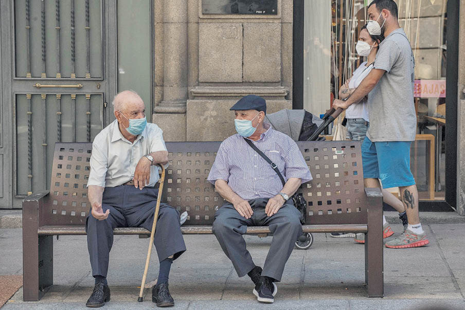 Dos personas mayores se protegen con mascarilla mientras conversan en un banco en la calle.