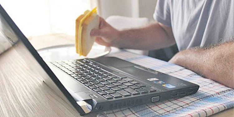 Un hombre come un sandwich delante de la pantalla de su ordenador portátil.